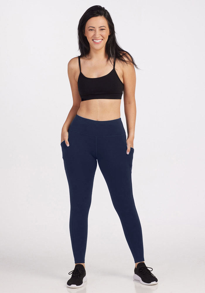 A woman smiles with her hands in the pockets of her Woolx Piper Pocket Leggings in Deep Navy, paired with a black sports bra and black sneakers, standing against a plain white background.
