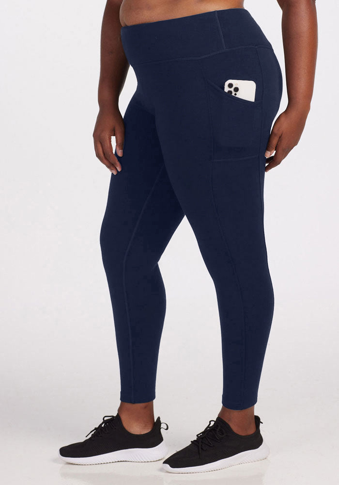 A person wearing Woolx Piper Pocket Tall leggings in Deep Navy, featuring a side pocket holding a phone, and black athletic shoes stands against a plain white background.