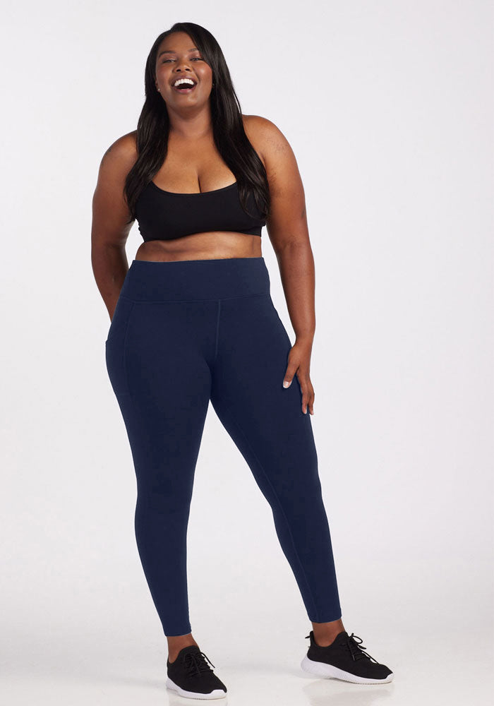 A woman with long dark hair smiles in Woolx’s Piper Pocket Tall high-waisted thermal leggings in Deep Navy, paired with a black sports bra and sneakers. She poses against a plain white background with one hand on her hip.