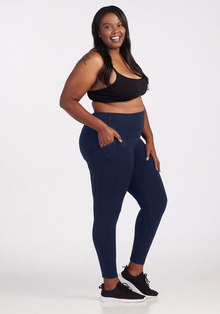 A woman smiles confidently in Woolx's Piper Pocket Leggings in Deep Navy, paired with a black sports bra and sneakers. She highlights the leggings’ convenient pockets against a plain white background.