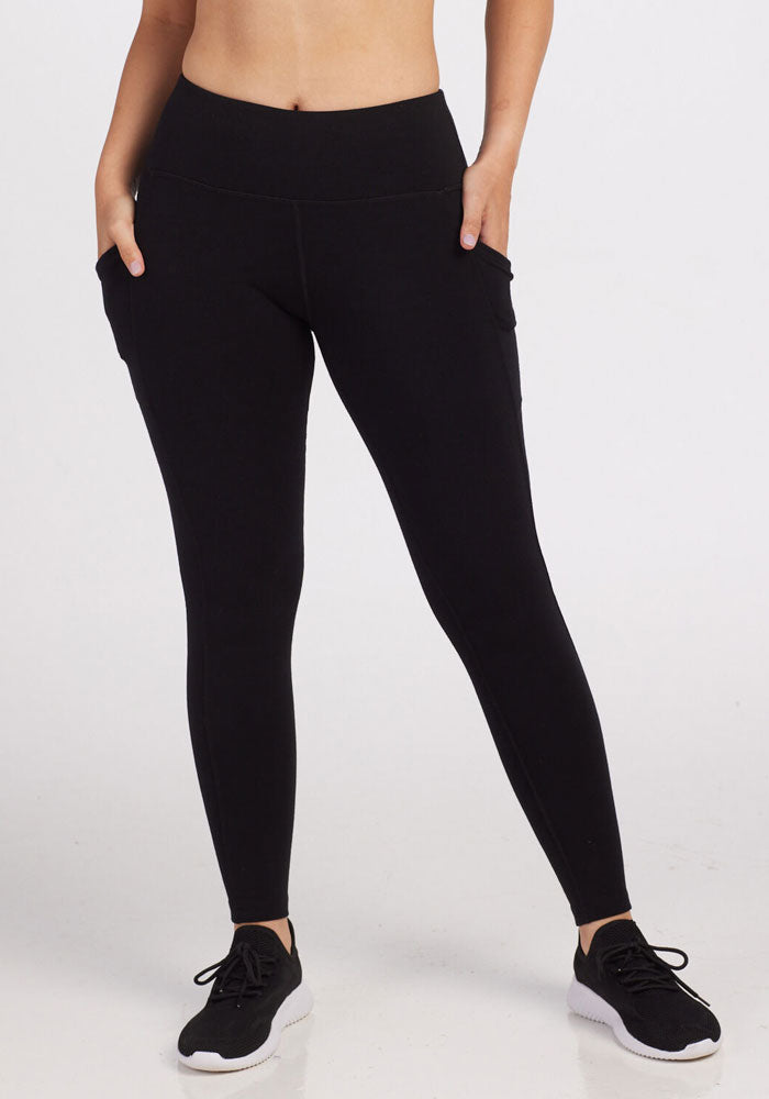 A person wearing Woolx's Piper Pocket Leggings in black, paired with sleek black sneakers, stands against a plain white background. Their hands are casually placed in the side pockets, highlighting the fit and style of these leggings.