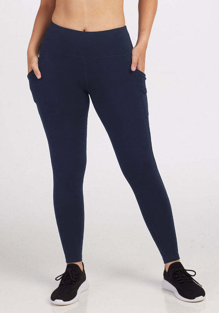 A person wearing Woolx Piper Pocket Leggings in Deep Navy, crafted from soft DuraLite fabric and featuring pockets, stands with hands in pockets. Black athletic shoes are worn; only the body from midriff to feet is visible against a plain background. 