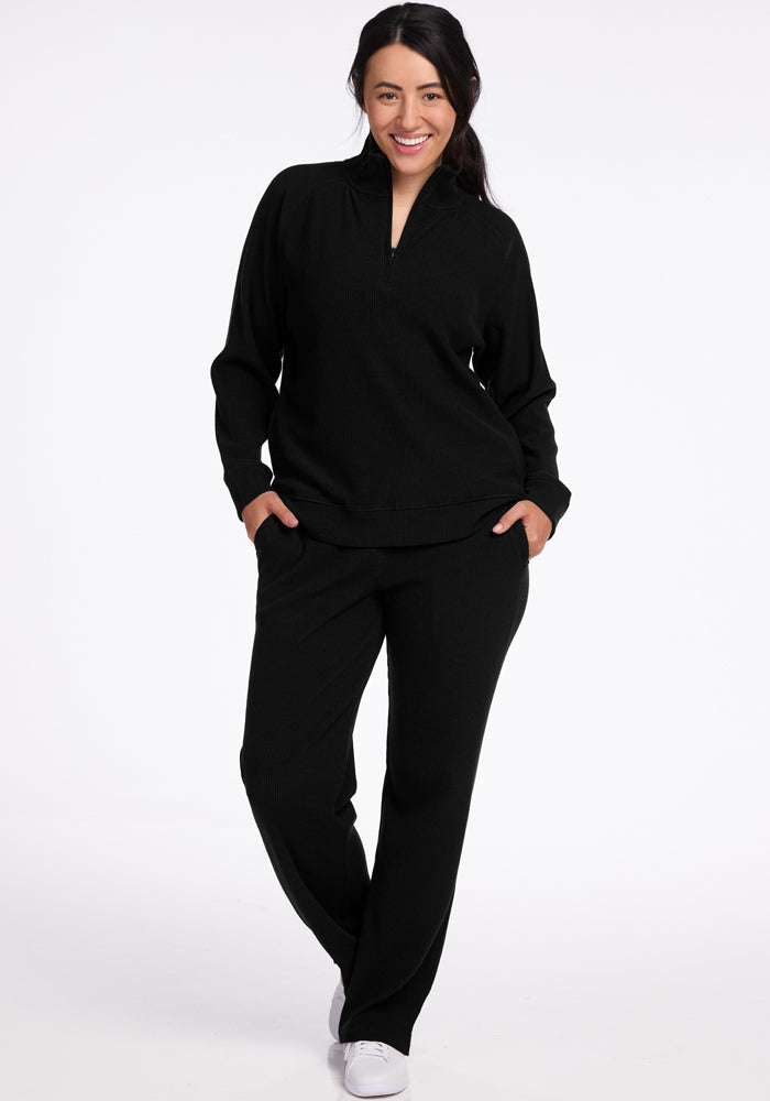 Smiling woman in Woolx Luca Petite Pants - Black, hands in pockets, stands against a white background.