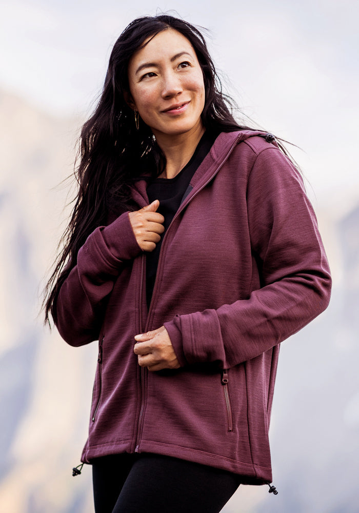 A woman with long dark hair wears the Woolx Cubby Hooded Sweatshirt in Grape Harvest over a black shirt, standing outdoors with blurred mountains behind her. She smiles and looks slightly to the side.