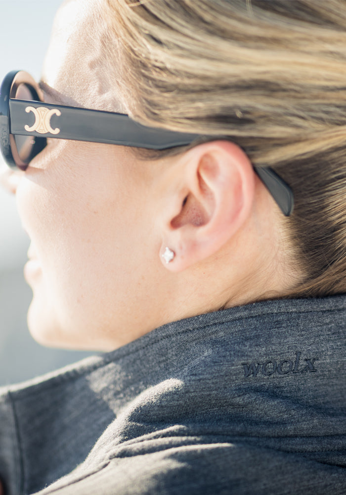 A close-up side view of a person in dark sunglasses and a Finley Zip Up Sweatshirt - Cinder Melange by Woolx, with "woolx" on the collar. Their blonde hair is pulled back, highlighting the cozy, odor-resistant Merino wool fabric.