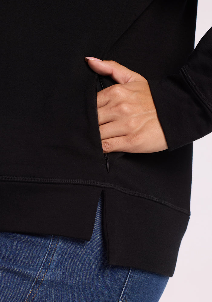 Wearing blue jeans and the Woolx Annie 3 Button Pullover in black, a person places their hand in a zippered side pocket; the pullover features a visible side seam and a small slit at the hem.