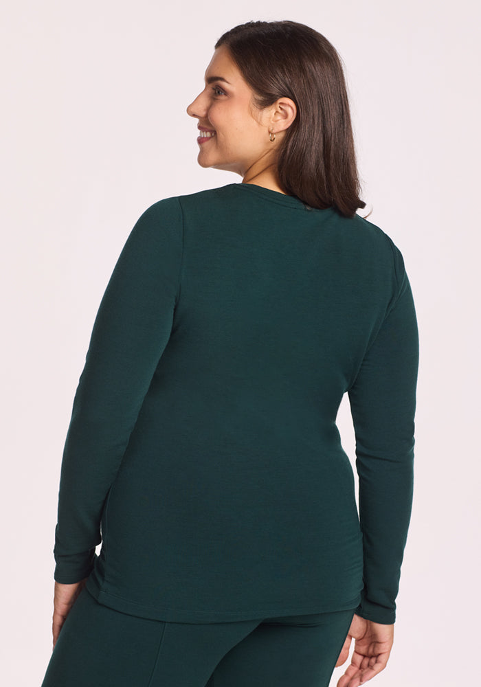 A woman with medium-length brown hair smiles while looking to the side, shown from the back against a light background. She wears the Woolx Sadie Baselayer Top in Deep Timber, a long-sleeve women's thermal made from Duralite merino fabric.
