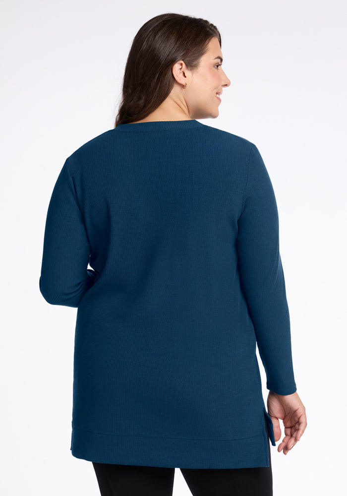 A woman with long brown hair, wearing the Woolx Evie Ribbed Tunic in Majolica Blue, stands facing away from the camera against a white background.