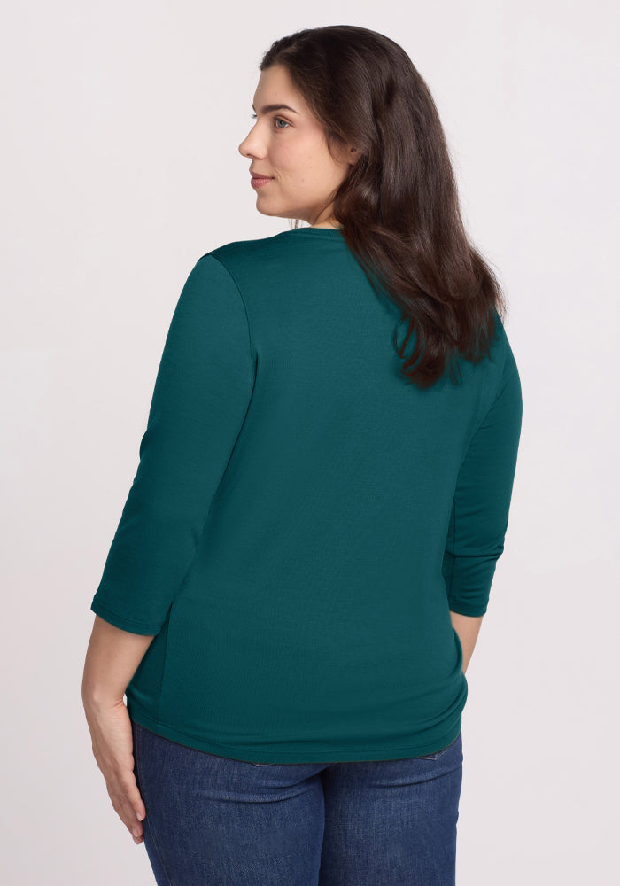A woman with long brown hair, wearing the Woolx Jenny 3/4 Sleeve Crew Neck in Riverbank Green and blue jeans, faces away from the camera looking over her left shoulder against a plain light background.