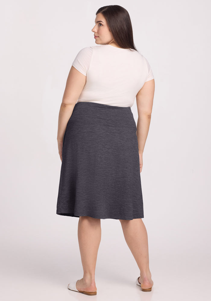 A woman with long brown hair stands facing away, wearing a short-sleeve white top, white flats, and the Woolx Marilyn Flowy Midi Skirt in Cinder Melange against a plain light background.
