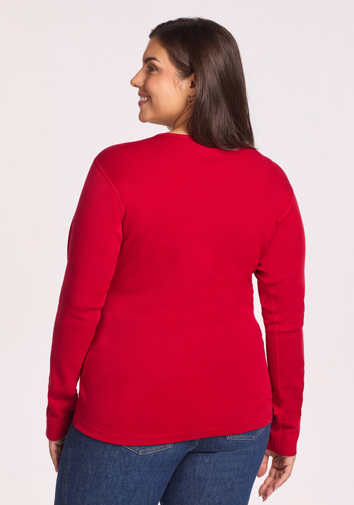 A woman with long dark hair smiles over her shoulder, wearing the Woolx Hannah Long Sleeve Top in Maraschino Cherry and blue jeans, standing against a plain light background.
