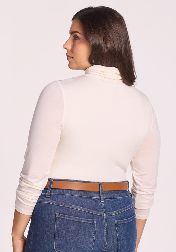 A woman with shoulder-length brown hair is shown from the back and side, wearing the Woolx Kennedy Bodysuit in Buttercream and blue jeans with a brown belt, standing against a plain light background.
