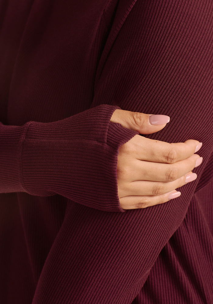 A close-up shows a person wearing the Woolx Ainsley Sweater in Toasted Garnet, featuring a ribbed design and thumb hole, with their hand resting on their upper arm and nails manicured in light pink.