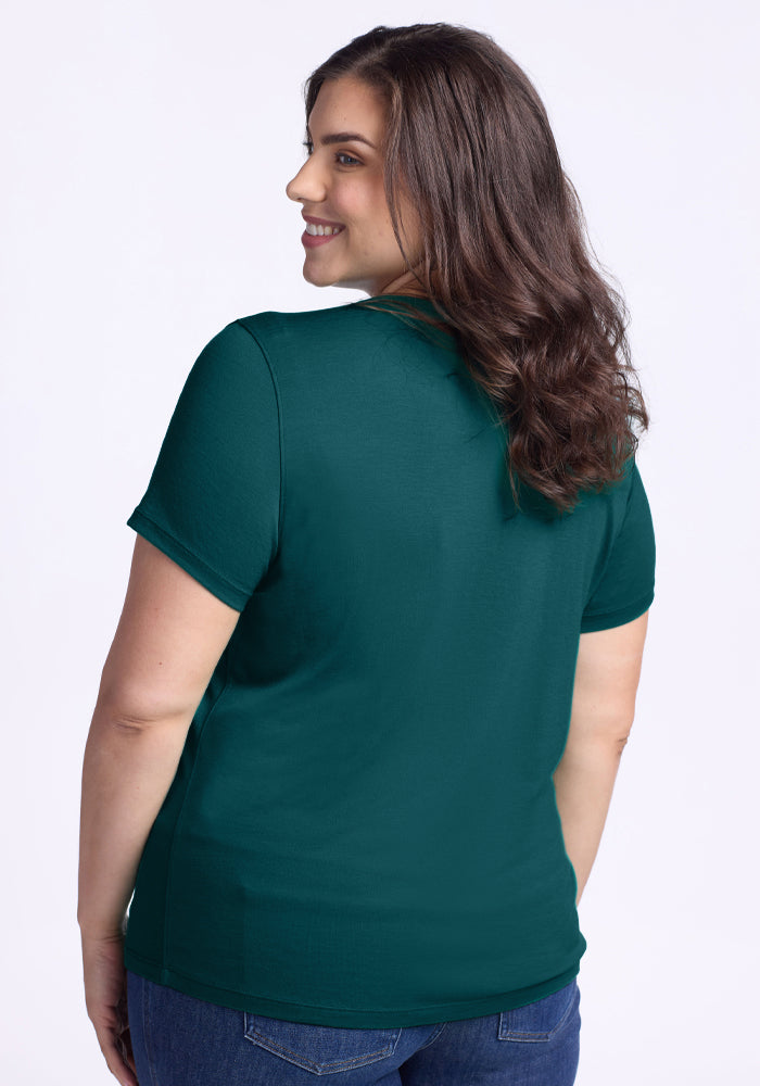 A woman with wavy brown hair wears the Woolx Liza Crew Neck Tee in Riverbank Green and blue jeans, standing against a light plain background, smiling as she turns her head to the side.