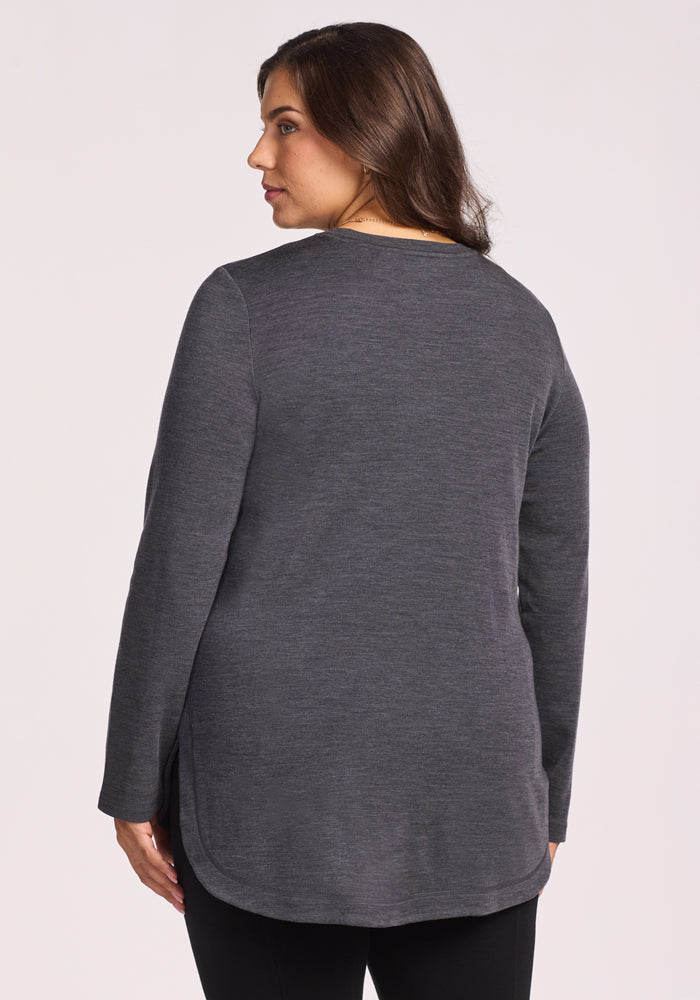 A woman with long brown hair is shown from the back, wearing the Woolx Blair Tunic in Cinder Melange—a long-sleeved Merino wool top paired with black pants—set against a plain light background.