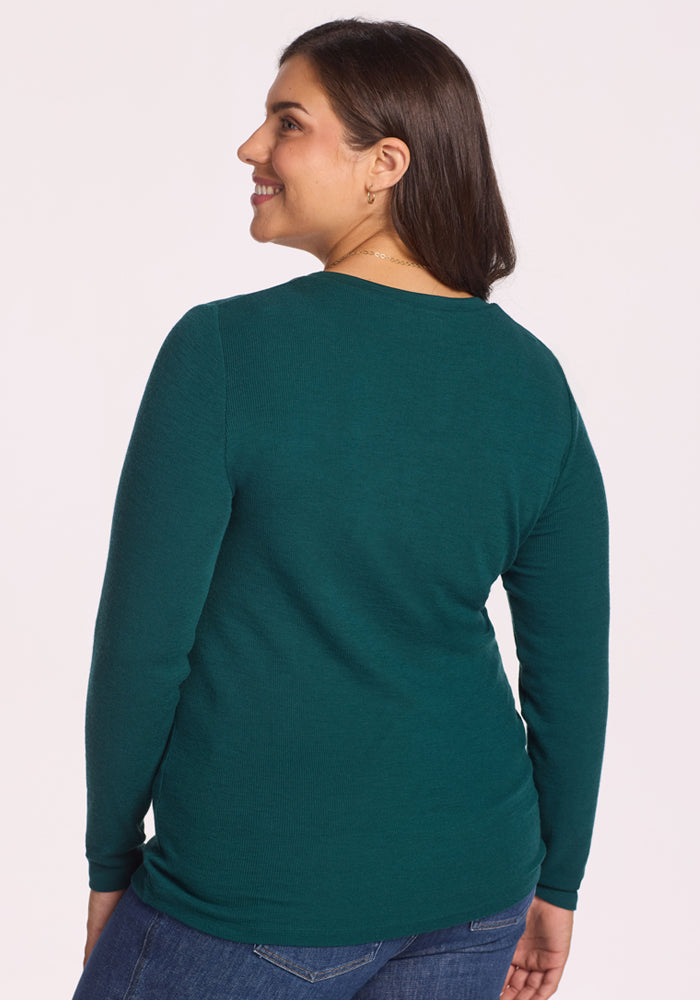A woman with long dark hair, wearing the Woolx Reese Ribbed Henley in Riverbank Green and blue jeans, smiles over her shoulder to showcase the back of her outfit against a plain light background.