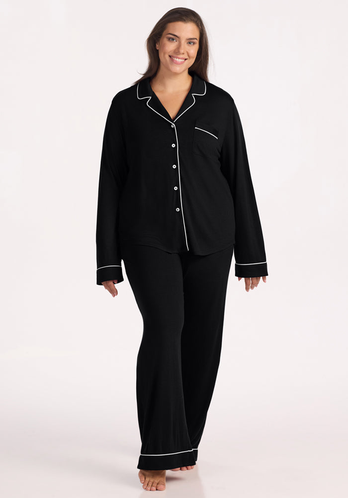 A woman with long brown hair smiles while wearing the Woolx Harper FeatherTouch® Pajama Set in black, featuring white trim, a button-up top with chest pocket, and matching pants. She stands barefoot against a plain light background. 