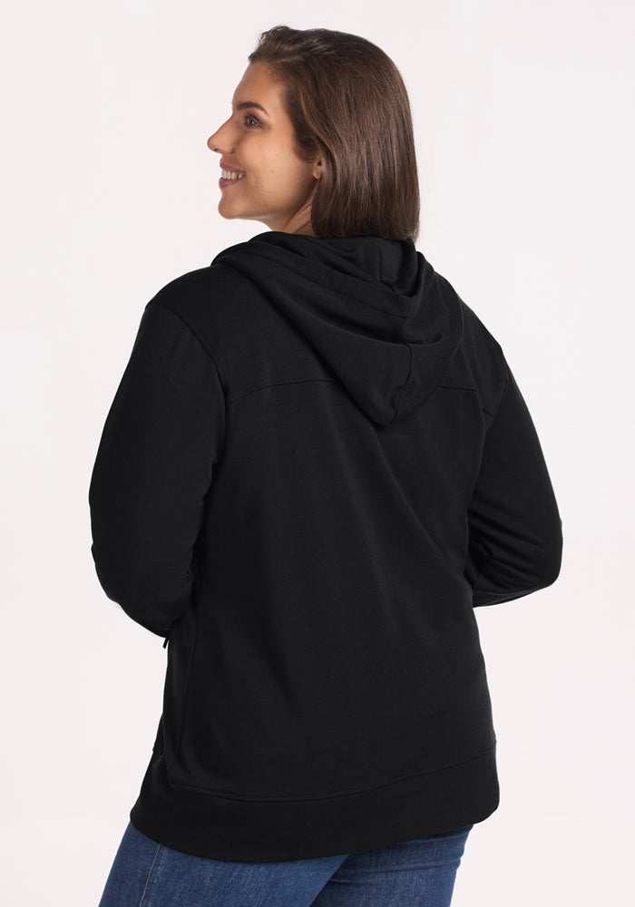 A woman with long brown hair, wearing the Woolx Darby Zip Up Hoodie in black and blue jeans, stands smiling and facing away from the camera against a plain light background.