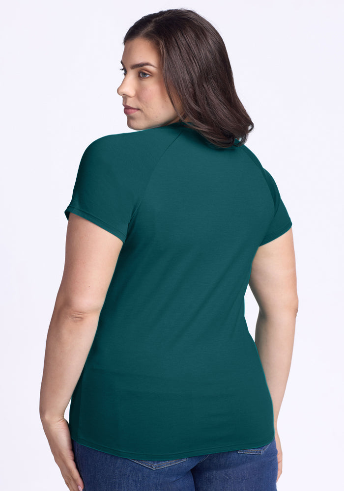 A woman with long brown hair, dressed in blue jeans and the Woolx Mia V Neck in Riverbank Green, stands facing away from the camera against a plain white background.