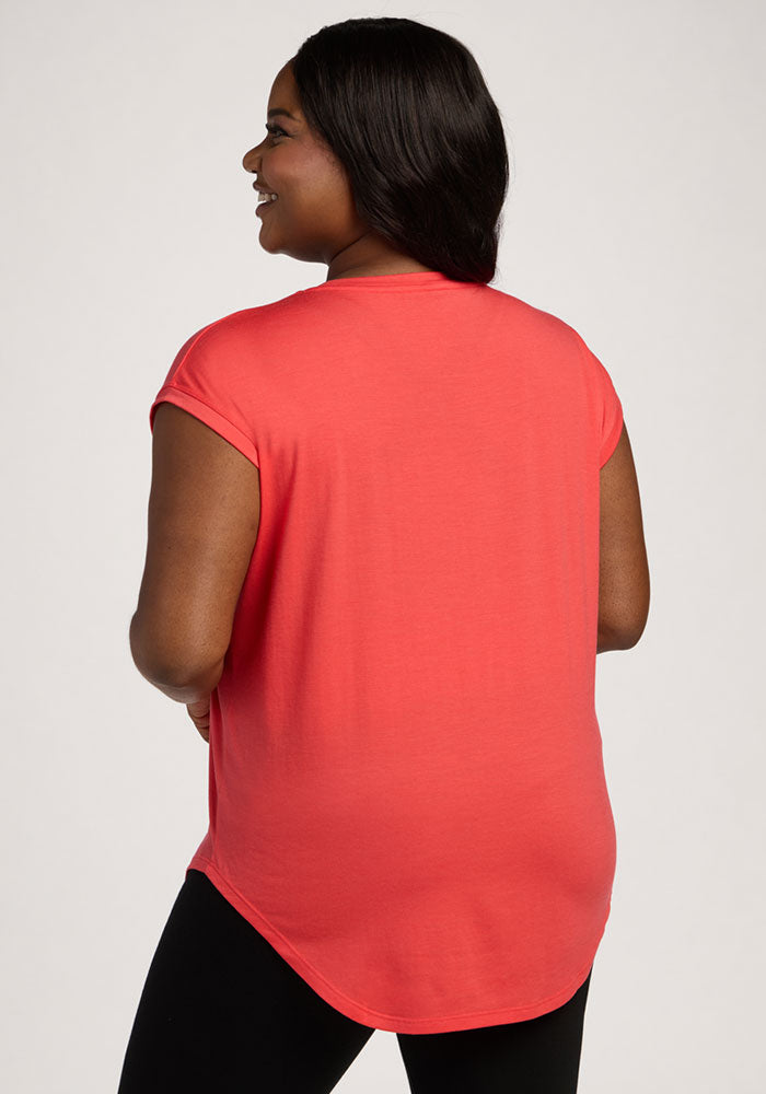 Woman faces away in Woolx Gabi Tunic Top - Hibiscus, short-sleeve coral, with black pants, on a plain background.