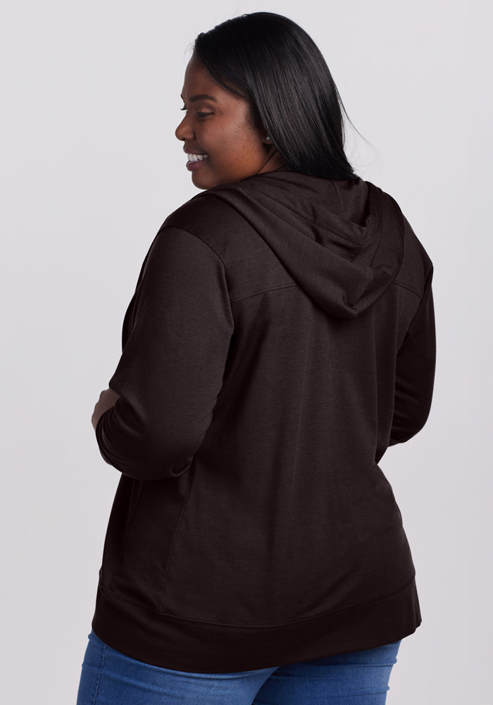 A woman with long dark hair, wearing the Woolx Ryann Hoodie in French Roast and blue jeans, stands smiling and facing slightly away from the camera against a plain light background.