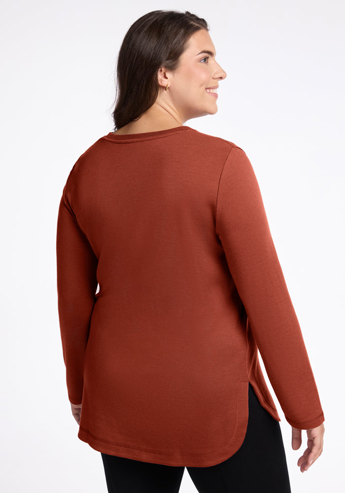 A woman with long brown hair smiles and faces away from the camera, wearing the Woolx Blair Tunic in Maple Leaf and black pants, set against a plain white background.