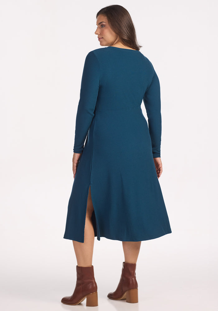 A woman faces away, wearing the Woolx Caroline Ribbed Twirl Dress in Majolica Blue with brown ankle boots. The merino wool's temperature-regulating fabric keeps her comfortable as she poses against a plain white background.