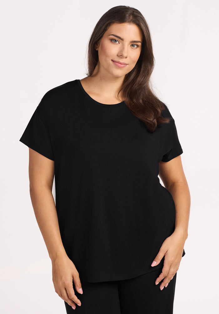 A woman with long brown hair, wearing the Woolx Billie Lounge Tee in black and matching pants, stands against a light background, smiling softly with one hand by her side.