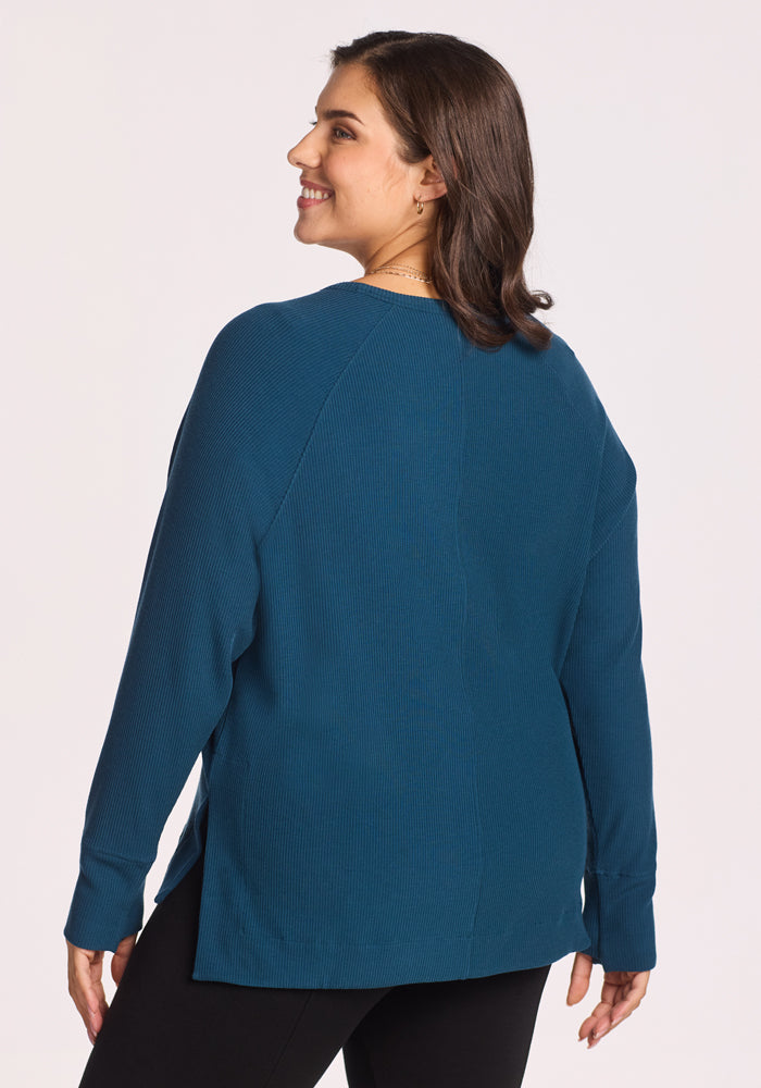 A woman with shoulder-length brown hair smiles while modeling the Woolx Ainsley Sweater in Majolica Blue. Shown from the back in black pants, this Merino wool sweater is both stylish and temperature-regulating.