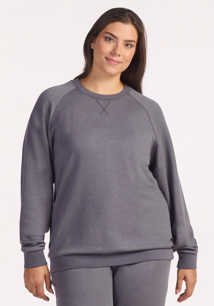 A woman with long brown hair wears the Woolx Bailey Crewneck Sweatshirt in Arctic Dusk and matching pants, standing against a plain light background, softly smiling at the camera. 