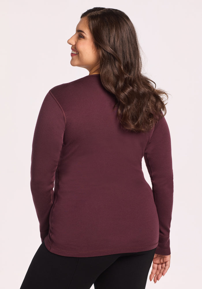 A woman with long brown hair stands facing away from the camera, wearing the Woolx Hannah Long Sleeve Top in Chocolate Fig and black pants. The temperature-regulating merino wool keeps her comfortable against a plain light background.