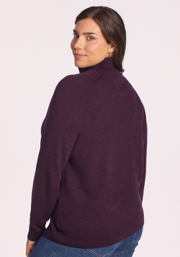 A woman with brown hair wears the Woolx Collins Half Zip Sweater in Deep Plum and blue jeans, standing slightly turned to the side and smiling gently over her shoulder against a light background.