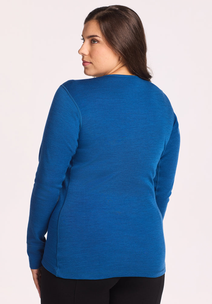 A woman with long brown hair stands facing away from the camera, wearing the Woolx Hannah Long Sleeve Top in Nightfall and black pants against a plain, light background.