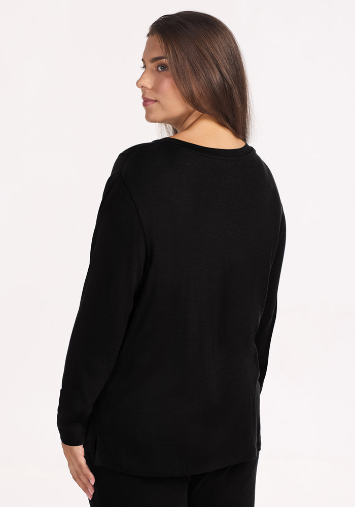 A woman with long brown hair models the Woolx Ashlie Lounge Top in black, a temperature-regulating Merino wool travel shirt. She stands against a plain white background, looking over her shoulder at the camera.