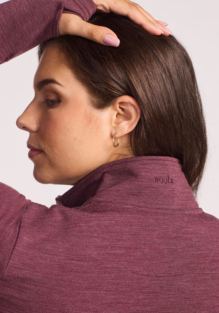 A woman with brown hair and a gold hoop earring wears the Woolx Finley Zip Up Sweatshirt in Grape Harvest Melange, featuring odor-resistant Merino wool. She rests one hand on her head, her profile softly lit.