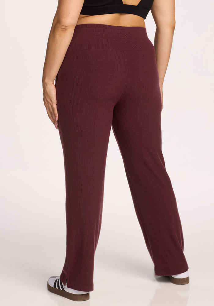 A person wearing Woolx Luca Ribbed Pants in Toasted Garnet and a black crop top stands with their back to the camera, also wearing black and white slide sandals.