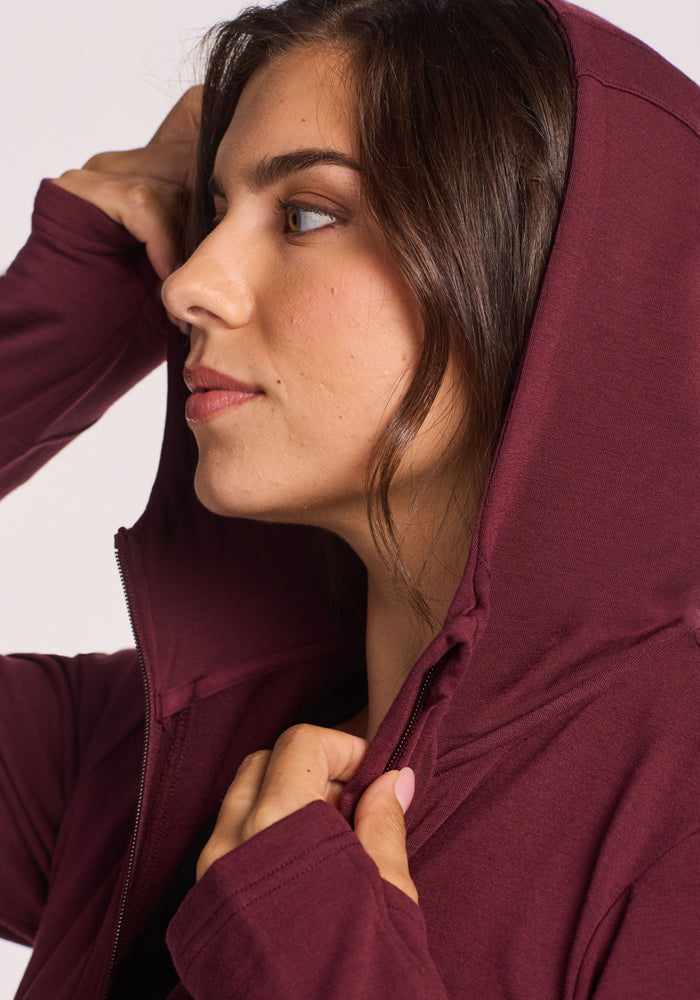 A woman with long brown hair wears a Woolx Zoey Zip Up Hoodie in Toasted Garnet, looking to the side. She holds the hood with one hand, her face partially framed by it against a light, plain background.