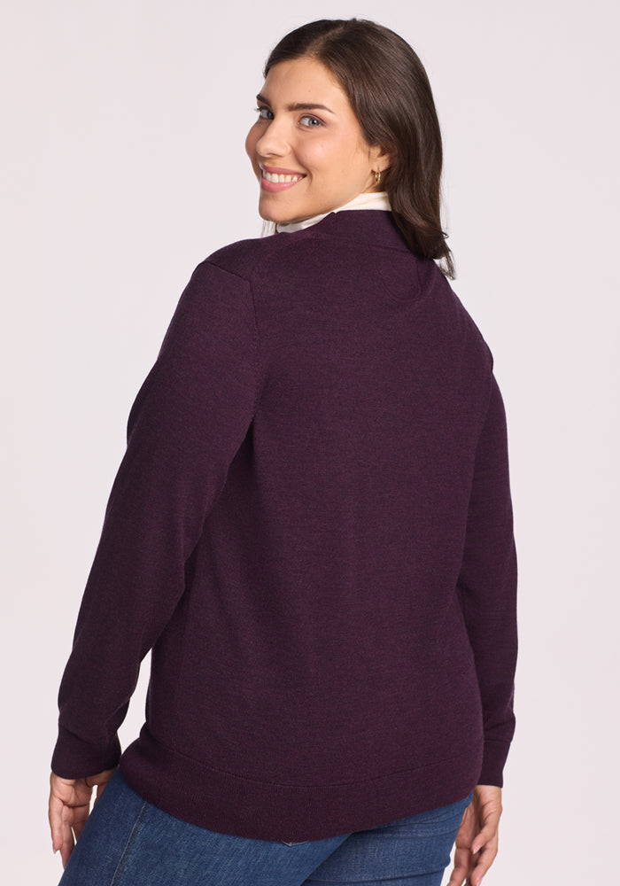 A woman with medium-length brown hair smiles over her shoulder, wearing the Woolx Gia Sweater in Deep Plum with blue jeans, standing against a plain light background.