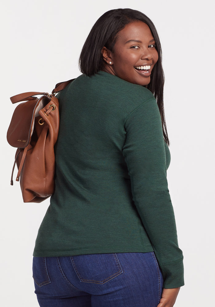 A smiling woman with long dark hair, wearing the Woolx Hannah Long Sleeve Top in Forest and blue jeans, carries a brown leather backpack over her shoulder against a plain white background.