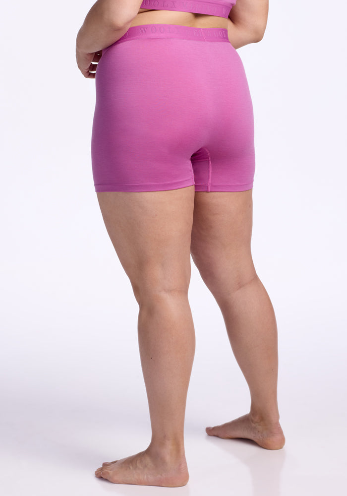 A person stands barefoot on a white surface, wearing a matching pink sports bra and Woolx Elliott Boyshort - Wild Blossom. The image is taken from behind, showing the person's legs and lower back.