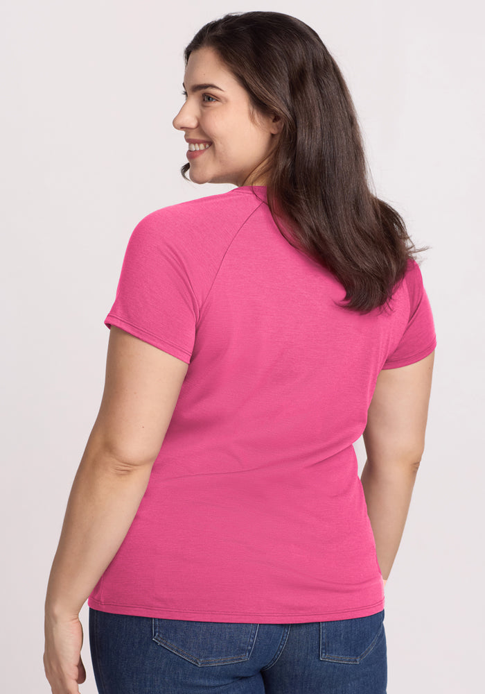 A woman with long brown hair, dressed in the Woolx Mia V Neck in Raspberry Fizz and blue jeans, smiles as she looks over her shoulder against a plain light background.