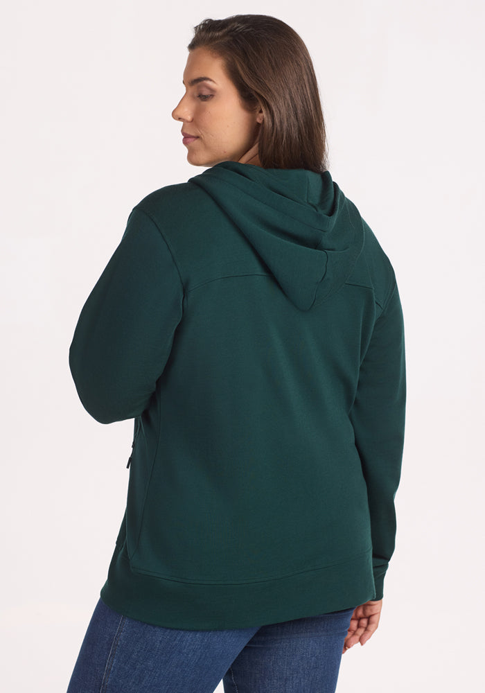 A woman with long brown hair models the Woolx Darby Zip Up Hoodie in Deep Timber, paired with blue jeans. Shown from the back, she looks over her shoulder against a plain white background.