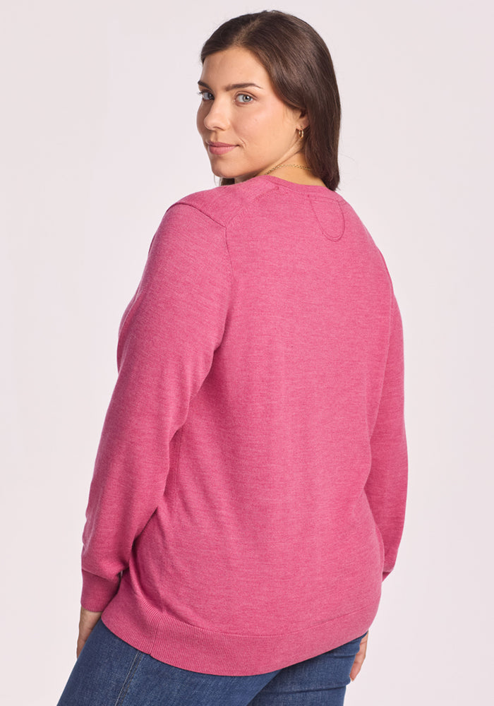 A woman with long brown hair stands against a plain light background, looking over her shoulder at the camera, wearing blue jeans and the Woolx Francesca Sweater in Wild Salmon.