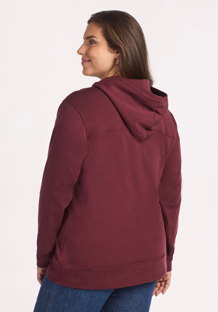 A woman with long brown hair, facing away, wears the Woolx Darby Zip Up Hoodie in Toasted Garnet and blue jeans against a plain white background.