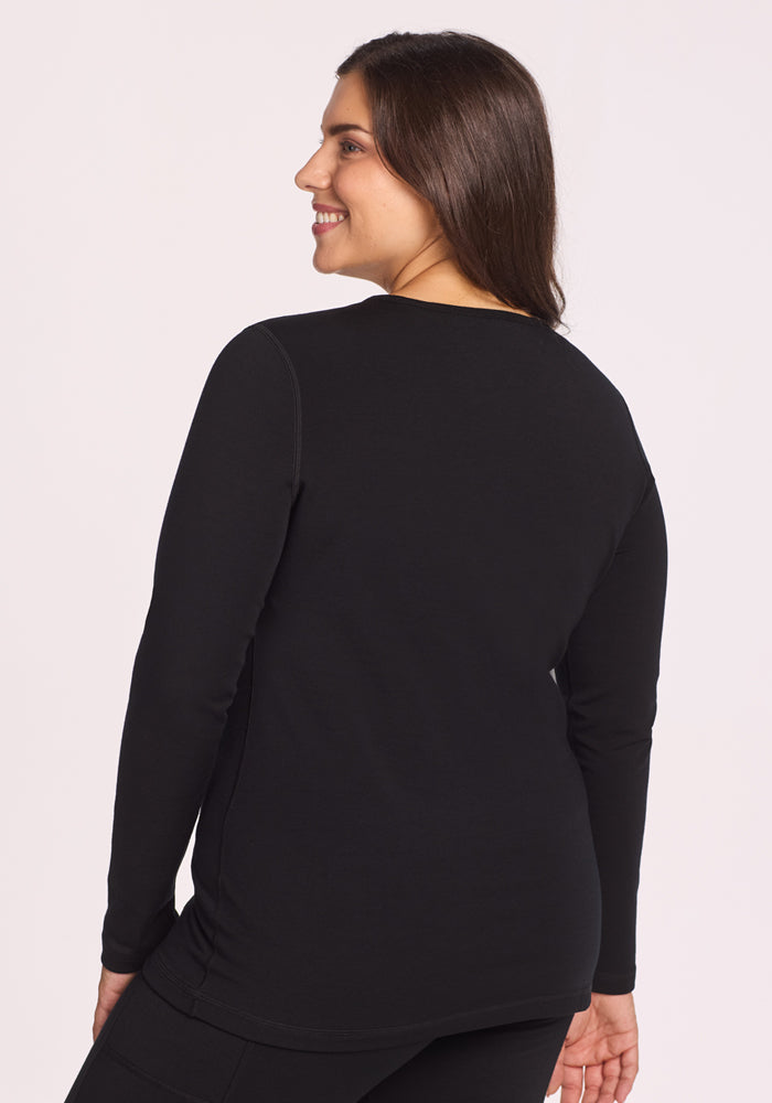 A woman with long brown hair smiles over her shoulder, wearing the Woolx Elsa Long Sleeve - Black top and black pants, standing against a plain light background.