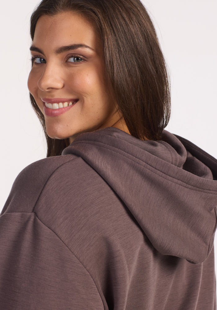 A young woman with long brown hair smiles over her shoulder, wearing the Woolx Avery Hoodie in Plum Truffle. The background is plain and light-colored.