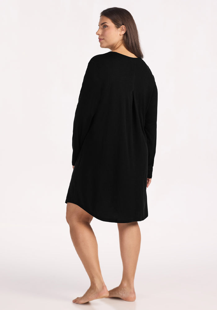 A woman with long brown hair stands barefoot, facing slightly to the side, wearing the Woolx Lucille Long Sleeve Nightgown in black. The plain light background highlights this cozy merino wool sleepwear choice.