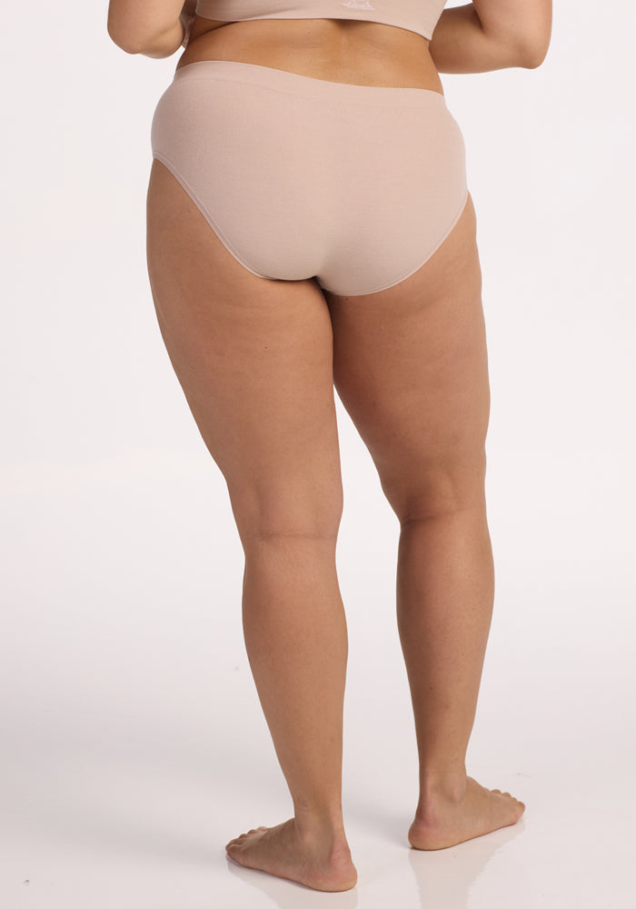 A person stands barefoot on a white background, shown from the waist down and back, wearing the Woolx Maeve Bikini in Sand, seamless moisture-wicking underwear with a matching bra.