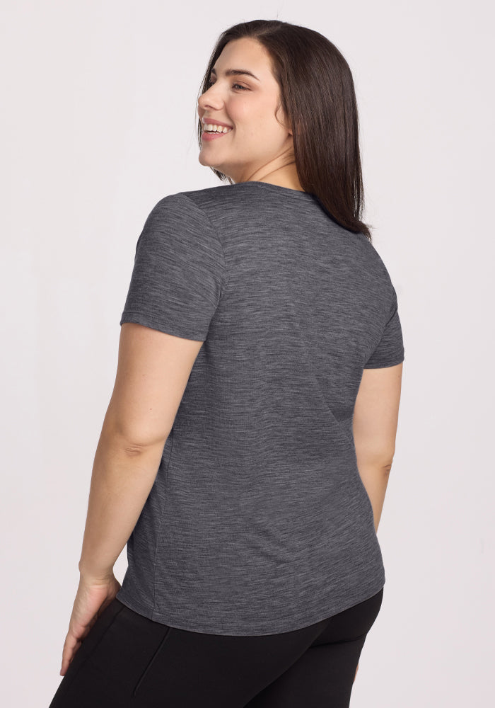 A woman with long dark hair smiles over her shoulder, facing away from the camera. She wears the Woolx Junie V Neck in Cinder Melange, a women's relaxed fit heathered gray Merino wool tee, paired with black pants against a plain background.