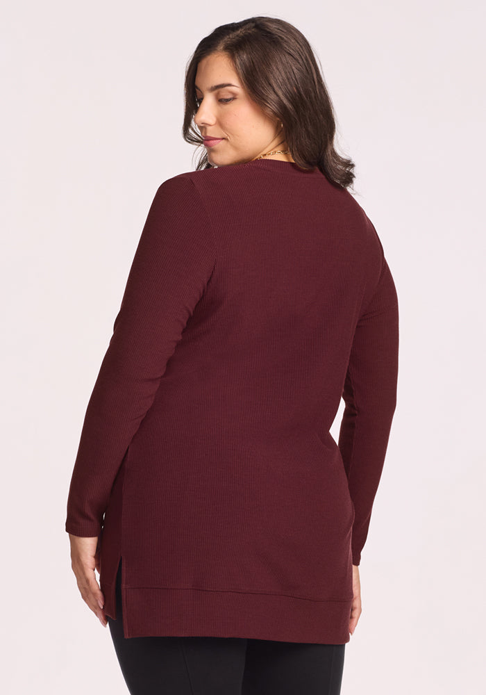 A woman with shoulder-length brown hair, seen from the back, wears the Woolx Evie Ribbed Tunic in Toasted Garnet with side slits and black pants, standing against a plain light background.